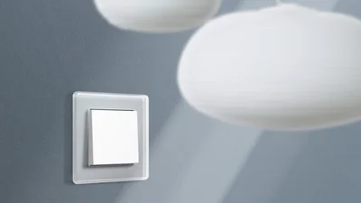 Ein moderner, weißer Lichtschalter mit einem Rahmen aus mattiertem Glas ist an einer grauen Wand montiert. Im unscharfen Hintergrund sind zwei helle, kugelförmige Lampen zu sehen, die eine sanfte Beleuchtung erzeugen.