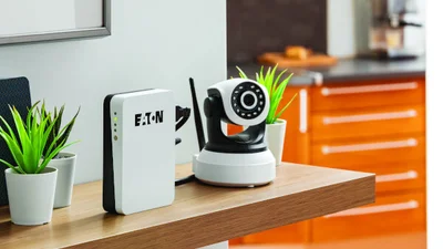Auf einem Regalboden aus Holz steht eine Eaton 3S Mini USV neben einer IP-Kamera. Links und rechts von Eaton 3S Mini USV und Kamera stehen kleine Pflanzen in weißen Übertöpfen. Im Hintergrund ist ein Schubladenschrank in orange erkennbar.