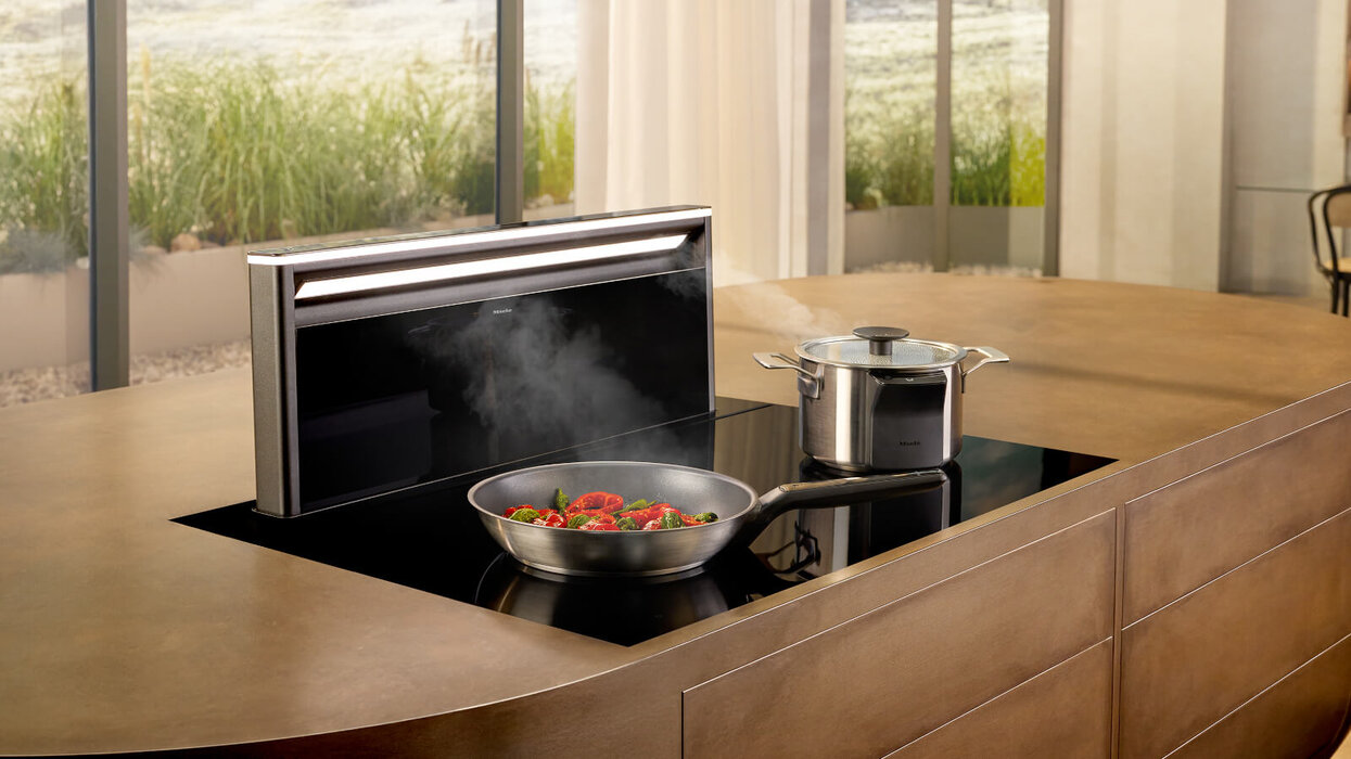 Das Bild zeigt eine moderne Küchensituation mit einer Kochinsel, einem schwarzen Kochfeld, einer Pfanne mit Gemüse, einem Topf und einer versenkbaren Dunstabzugshaube im Hintergrund.