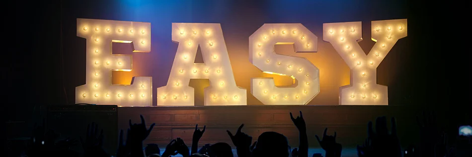 Das Bild zeigt große, beleuchtete Buchstaben, die das Wort "EASY" auf einer Bühne darstellen. Im Vordergrund sind Silhouetten von jubelnden Menschen zu sehen.