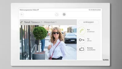 Eine weiße Gira Wohnungsstation Video IP zeigt die Video-Türkommunikation mit Bildvorschau und Bedienelementen wie Ruf annehmen und Tür öffnen. Im Display ist eine Person vor dem Hauseingang zu sehen.
