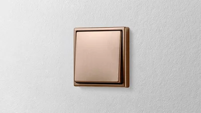 Das Bild zeigt einen bronzefarbenen Lichtschalter des Herstellers JUNG an einer weißen Wand.