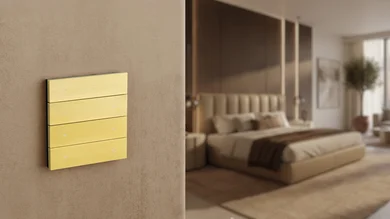 Ein messingfarbener Theben iONprime-Schalter ist an einer Wand im Schlafzimmer montiert. Im Hintergrund ist ein modern eingerichteter Raum mit Bett und warmen Holzelementen zu sehen.