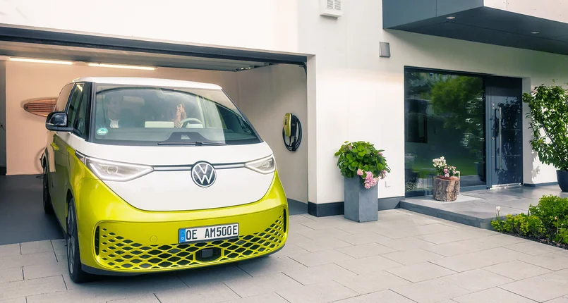 Das Bild zeigt einen modernen VW ID. Buzz, der aus einer Garage eines Einfamilienhauses fährt. An der Garagenwand ist eine Wallbox installiert. Die Umgebung wirkt gepflegt und modern, mit Pflanzen und klarer Architektur.