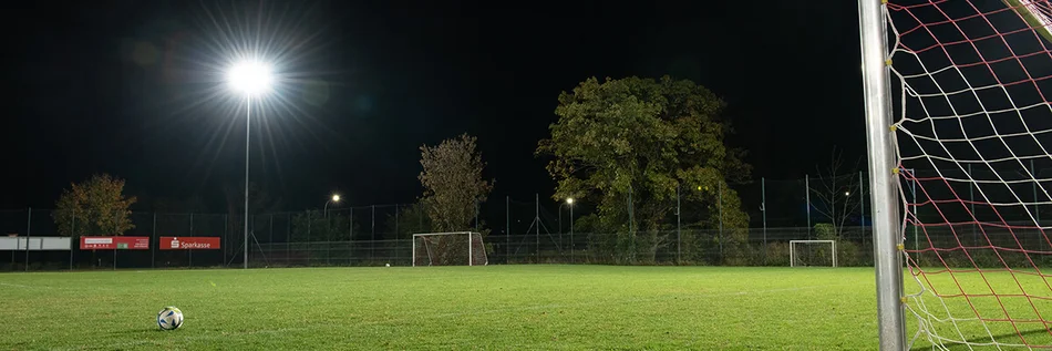 Das Bild zeigt einen Teil eine Fußballfeldes. Im Vordergrund ist rechts ein Teil des Tores sichtbar. Der Fußballplatz wird durch einen Fluter beleuchtet, der in einer Ecke des Platzes steht. 