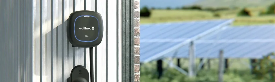 Das Bild zeigt eine Wallbox-Ladestation an einer Metallwand, im Hintergrund sind Solarpanels auf einer Wiese zu sehen.