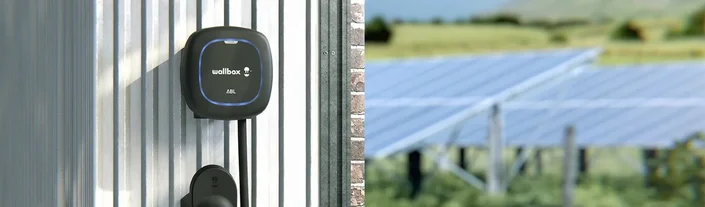 Das Bild zeigt eine Wallbox-Ladestation an einer Metallwand, im Hintergrund sind Solarpanels auf einer Wiese zu sehen.