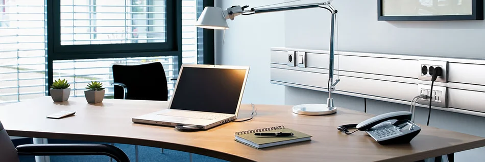 Arbeitsplatz mit Schreibtisch, Laptop, Stehlampe und Telefon. Im Hintergrund ein Leitführungsystem von OBO Bettermann.
