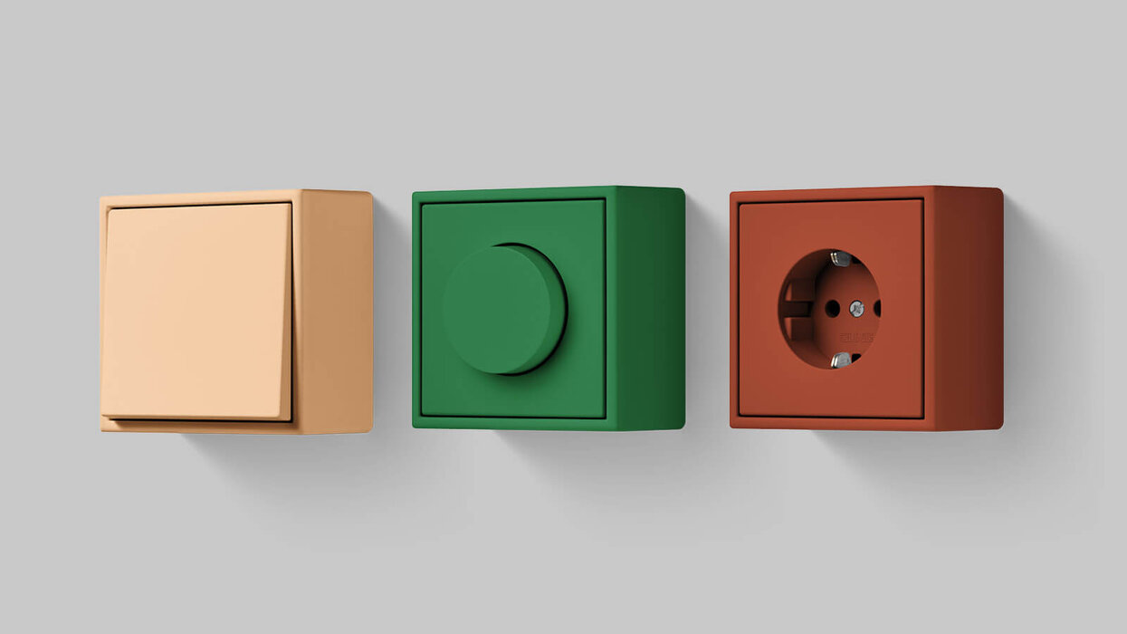 3 Aufputz Schalter und Steckdosen in verschiedenen Farben in verschiedenen Farben. Links Schalter in der Farbe Beige. Mitte Drehschalter in der Farbe Grün. Rechts Steckdose in der Farbe Braun.
