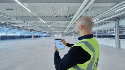 Eine Person mit gelber Warnweste steht in einer sehr großen Halle und bedient das Display seines Handys.