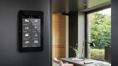 Zu sehen ist ein schwarzes KNX-Smart-Panel an der Wand in einem modern eingerichteten Raum. Auf dem Display werden verschiedene Wohnbereiche und Temperaturen angezeigt.