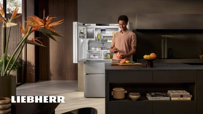Das Bild zeigt eine moderne Küche, in der eine Person vor einem geöffneten Liebherr-Kühlschrank steht und ein Getränk zubereitet.