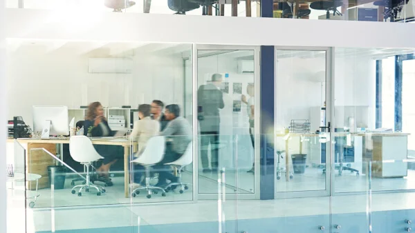 Das Bild zeigt ein modernes, helles Großraumbüro mit Glaswänden, in dem mehrere Personen an einem Tisch sitzen und sich in einem Meeting unterhalten.