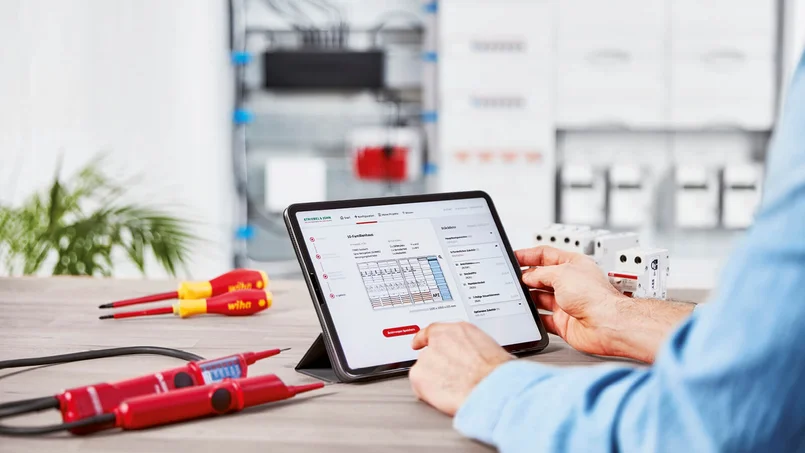 Eine Person sitzt an einem Tisch und bedient ein Tablet. Auf dem Tisch liegen Messgeräte, Schraubendreher und mehrere elektrische Bauteile, im Hintergrund ist ein Schaltschrank zu sehen.