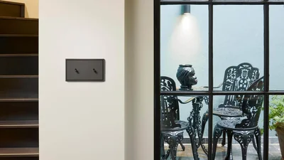 Das Bild zeigt einen modernen Doppeltoggleschalter in dunklem Design an einer hellen Wand neben einer Treppe. Durch das Fenster ist ein stilvoller Außenbereich mit schwarzen Metallstühlen und einem Tisch sichtbar.