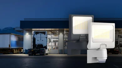 LED-Strahler mit integriertem Bewegungsmelder für die energieeffiziente Außenbeleuchtung von Gebäuden. Im Hintergrund ist ein beleuchteter Logistik- bzw. Werkstattbereich mit Lkw zu sehen.