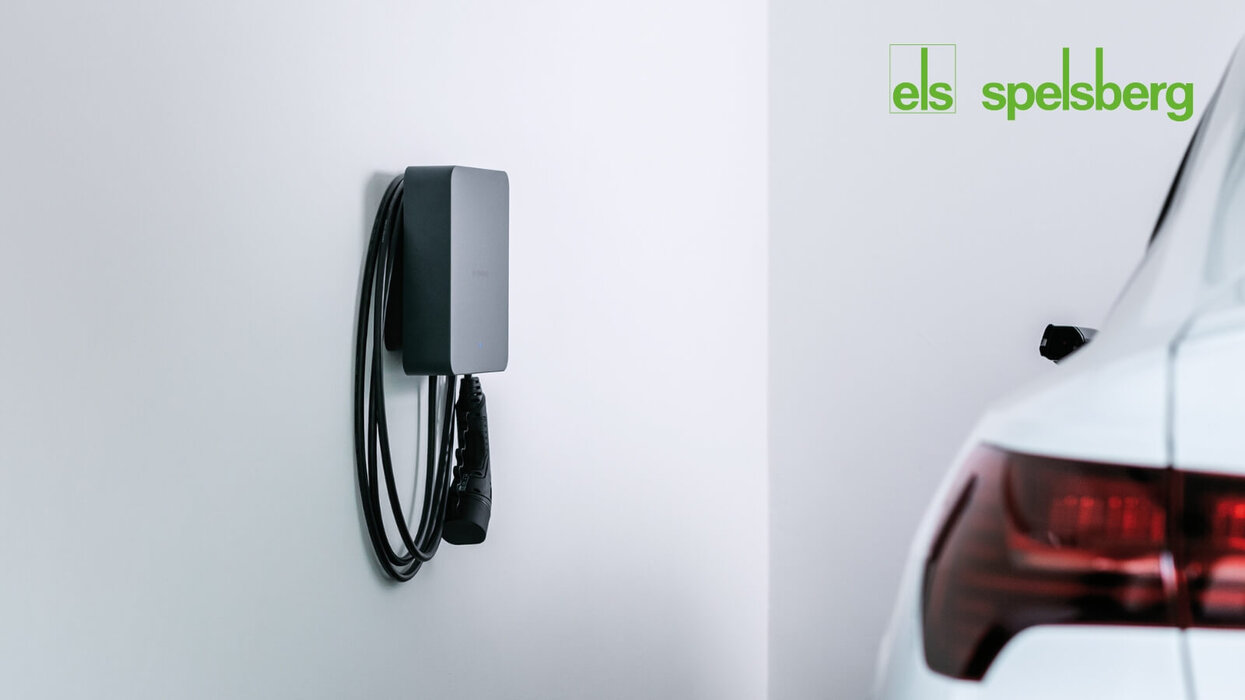 Das Bild zeigt eine moderne Wallbox zur Ladung eines Elektroautos, die an einer weißen Garagenwand montiert ist. Rechts ist das Heck eines weißen Fahrzeugs zu sehen, oben rechts befindet sich das Spelsberg-Logo.