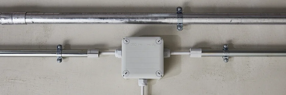 Verbindungs- und Befestigungssystem von OBO Bettermann an der wei0en Wand. In der Mitte ist ein kleiner weißer Kasten zu sehen sehen, von dem schmale Rohre nach rechts , links und unten abgehen.