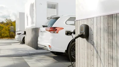 Das Bild zeigt eine Wallbox-Ladestation an einer Holzfassade, die ein weißes Elektroauto lädt. Im Hintergrund ist eine weitere Ladestation mit einem Fahrzeug zu sehen.
