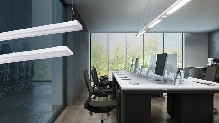 Das Bild zeigt ein modernes Großraumbüro mit großen Fenstern und mehreren Arbeitsplätzen. Auffällig sind die schlanken, rechteckigen LED-Leuchten, die von der Decke abgehängt sind und für eine gleichmäßige, helle Ausleuchtung des Raumes sorgen. Die Leuchten unterstreichen das zeitgemäße, minimalistische Design und schaffen eine angenehme Arbeitsatmosphäre.