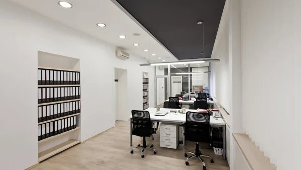 Das Bild zeigt einen weiß gestrichenen, länglichen Büroraum, der an der Stirnseite verglast ist. In die linke Wand ist eine Regalnische eingelassen, die mit vielen schwarzen Aktenordnern gefüllt ist. Auf der rechten Raumseite stehen vier große Schreibtische hintereinander. An jedem Schreibtisch stehen 2 Bürostühle nebeneinander. Licht spenden sowohl längliche Hängeleuchten über den Schreibtischen als auch Downlights an der Decke.