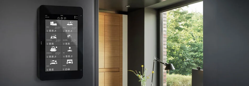 Zu sehen ist ein schwarzes KNX-Smart-Panel an der Wand in einem modern eingerichteten Raum. Auf dem Display werden verschiedene Wohnbereiche und Temperaturen angezeigt.