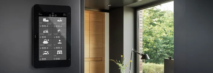 Zu sehen ist ein schwarzes KNX-Smart-Panel an der Wand in einem modern eingerichteten Raum. Auf dem Display werden verschiedene Wohnbereiche und Temperaturen angezeigt.