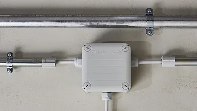 Verbindungs- und Befestigungssystem von OBO Bettermann an der wei0en Wand. In der Mitte ist ein kleiner weißer Kasten zu sehen sehen, von dem schmale Rohre nach rechts , links und unten abgehen.
