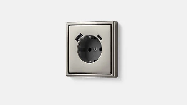 Einzelansicht einer Steckdose mit USB Anschlüssen des Herstellers Jung in der Farbe Silber vor einem hellen Hintergrund.