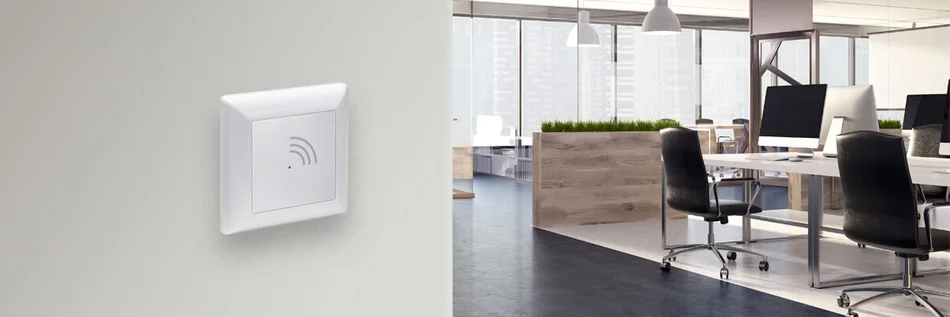 Das Bild zeigt einen Wandschalter mit RFID-Symbol in einem modernen, offenen Büro mit mehreren Arbeitsplätzen.