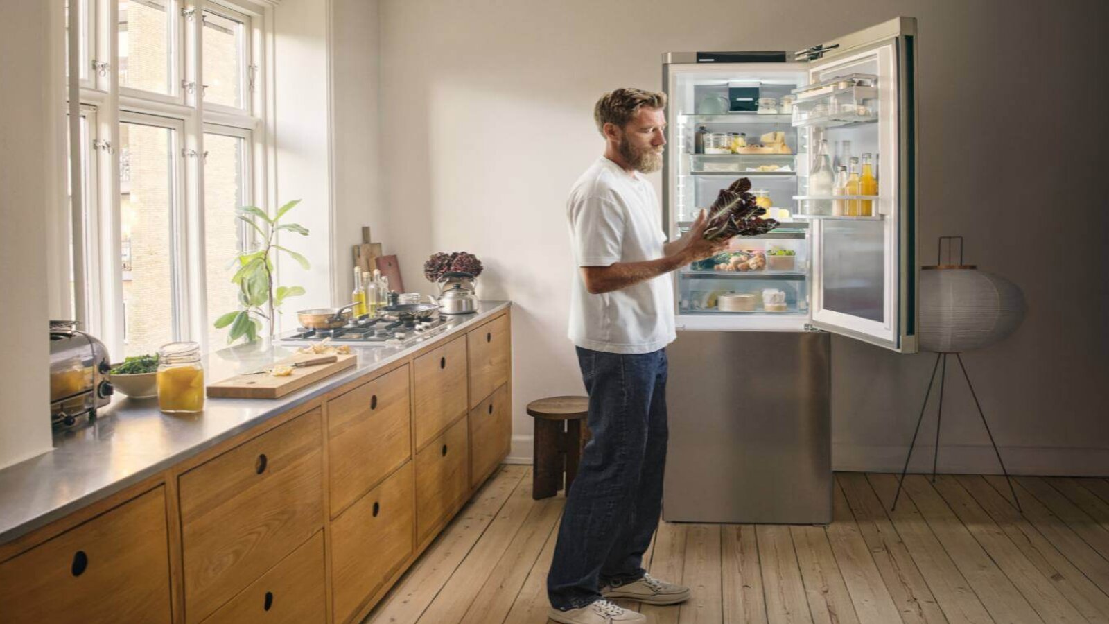 Eine Person steht in einer hellen Küche vor einem geöffneten Kühlschrank und nimmt frische Lebensmittel heraus.