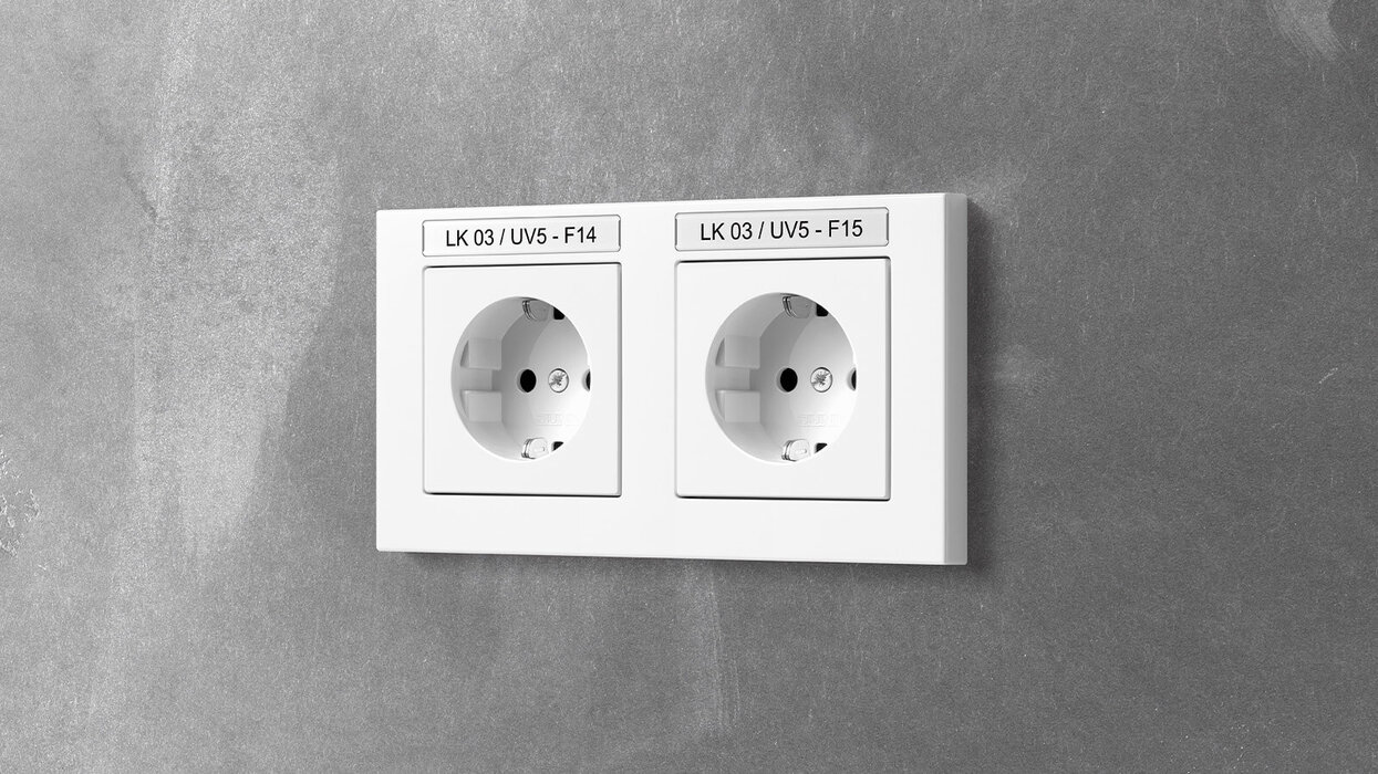Das Bild zeigt eine doppelte, weiße Steckdose mit Beschriftungsfeldern an einer grauen Wand.