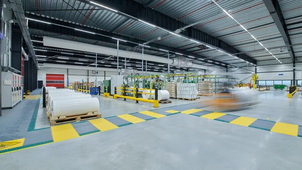 Das Bild zeigt eine moderne, helle Produktions- oder Lagerhalle mit klar strukturierter Einrichtung und Sicherheitsmarkierungen auf dem Boden. Im Vordergrund sind große weiße Rollen auf Paletten zu sehen, während im Hintergrund Maschinen, Regale mit Materialien und ein verschwommen vorbeifahrendes Flurförderfahrzeug (z. B. Gabelstapler) erkennbar sind. An der Decke verlaufen Lichtbänder. 