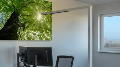 Das Bild zeigt einen Schreibtisch mit einem Bildschirm und davor stehendem Stuhl. Über dem Schreibtisch hängt ein großes Foto eines begrünten Baumes. Auf den Schreibtisch fällt Licht von der davorstehenden Büroleuchte STYLEbutler von Dotlux