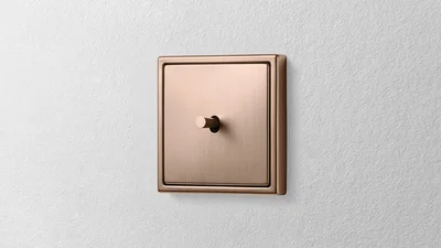 Das Bild zeigt einen bronzefarbenen, rechteckigen Schalter mit Kipphebel an einer weißen Wand