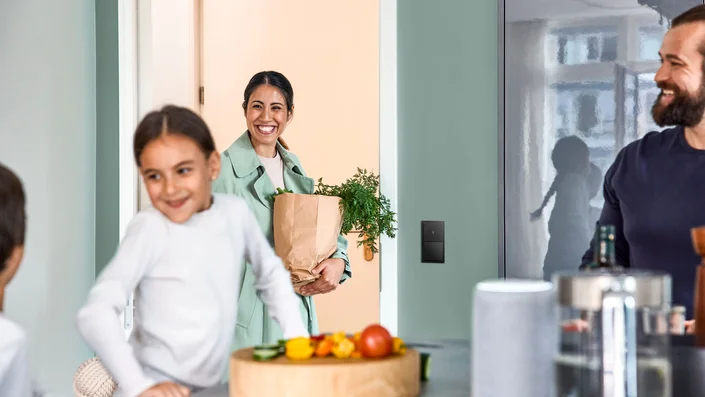 Auf dem Bild kommt eine Frau mit einer Einkaufstüte in eine moderne Küche, in der bereits Kinder und ein Mann sind. Gemüse und Haushaltsgegenstände sind zu sehen.