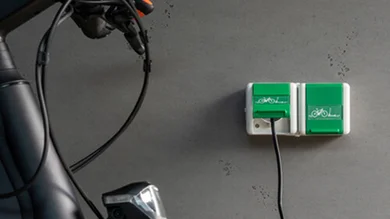 Zwei Steckdosen an der Wand montiert. Im Vordergrund ein Fahrrad. Steckdose mit einer Klappe in der Farbe Grün und einem Fahrradsymbol in der Farbe Weiß.