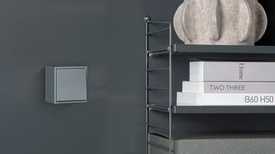 Aufputz Schalter in der Farbe Hellgrau an einer dunkelgrauen Wand montiert. Daneben ein Regal aus Metall mit Büchern .