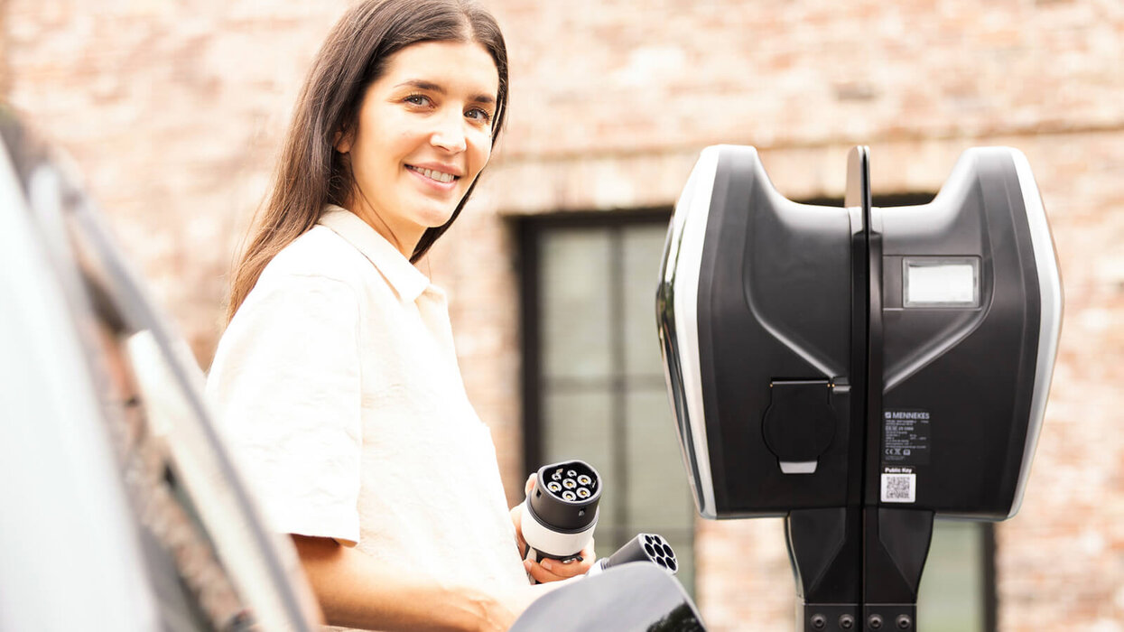 AMTRON® 4Business 700 von Mennekes Eine Frau steht neben einer schwarzen Ladestation für Elektroautos. Sie hält einen Ladestecker in der Hand und schaut in die Kamera. Die Ladestation befindet sich im Freien vor einer Backsteinwand mit einem Fenster.