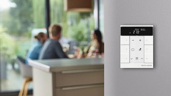 Das Bild zeigt ein weißes Bedienpanel mit Display und Tasten an einer Wand, im Hintergrund sitzen drei Personen an einem Tisch.