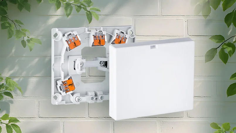 Das Bild zeigt eine geöffnete, weiße Herdanschlussdose mit integrierten Verbindungsklemmen an einer hellen Wand im Außenbereich. Umgeben ist die Szene von grünem Blattwerk.