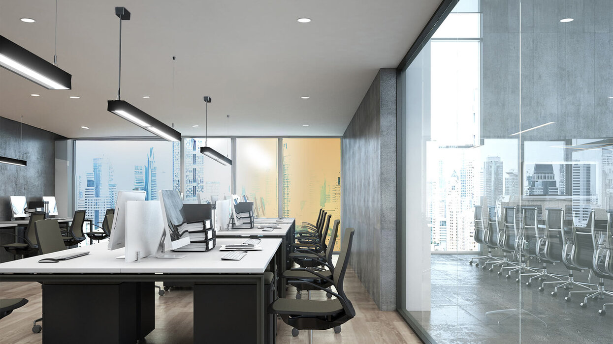 Das Bild zeigt ein modernes Open-Space-Büro mit Reihen von Arbeitsplätzen, großen Fensterfronten und schwarzen Pendelleuchten über den Tischen.