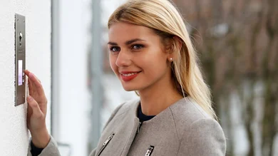 Das Bild zeigt eine Frau, die an einer Türsprechanlage im Außenbereich klingelt und dabei freundlich lächelt.