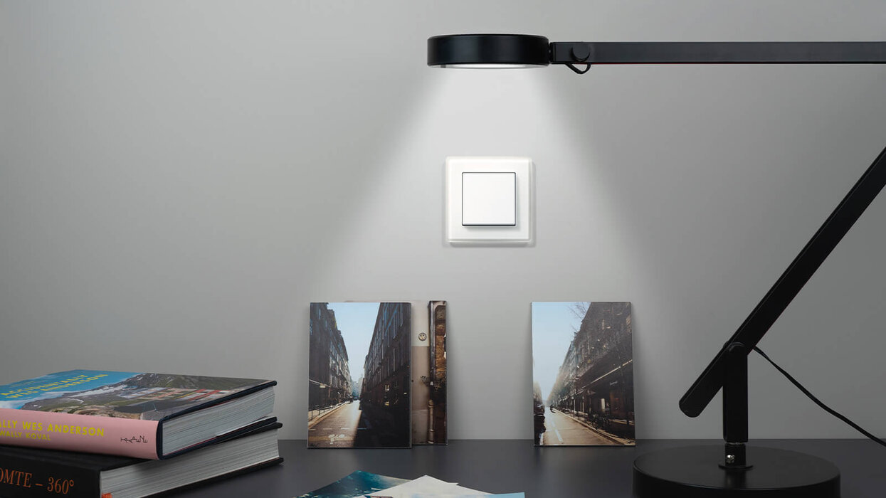 Schalter von JUNG im Hintergrund. Im Vordergrund ist eine moderne und angeschaltete Lampe zu sehen.