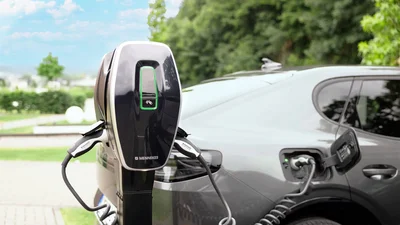 Ein schwarzes Elektroauto ist an eine schwarze Ladestation angeschlossen. Das Ladekabel ist mit dem Fahrzeug verbunden. Im Hintergrund befinden sich grüne Bäume, ein Gehweg und ein wolkiger Himmel.