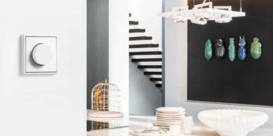 Weißer Drehdimmer an einer hellen Wand in einem modernen Wohnraum mit Esstisch, Treppe, Hängeleuchte und dekorativen Wandobjekten im Hintergrund.