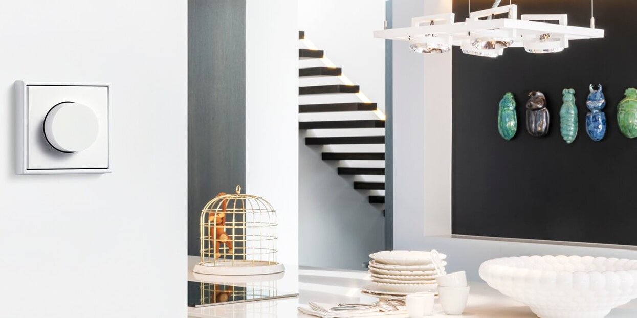 Weißer Drehdimmer an einer hellen Wand in einem modernen Wohnraum mit Esstisch, Treppe, Hängeleuchte und dekorativen Wandobjekten im Hintergrund.