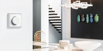 Weißer Drehdimmer an einer hellen Wand in einem modernen Wohnraum mit Esstisch, Treppe, Hängeleuchte und dekorativen Wandobjekten im Hintergrund.