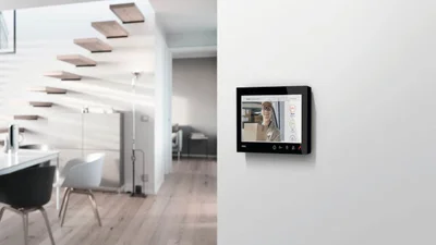 Das Bild zeigt im rechten Bildbereich eine schwarze Wohnungsstation an einer weißen Wand. Im rechten Bildbereich wird ein moderner Wohnbereich mit einem Essbereich gezeigt Eine freischwebende Treppe mit Holzstufen führt ins Obergeschoss.