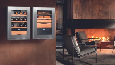 Dekorativer Weinkühlschrank an der Wand montiert mit Weinflaschen und im Hintergrund zwei elegante Sessel und Kaminfeuer.